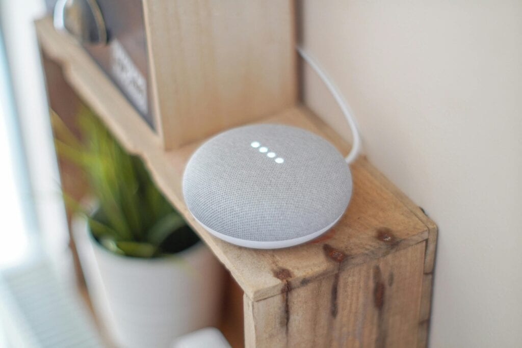 Google Nest Mini smart speaker in chalk grey sitting on a rustic wooden shelf with four white LED lights active.