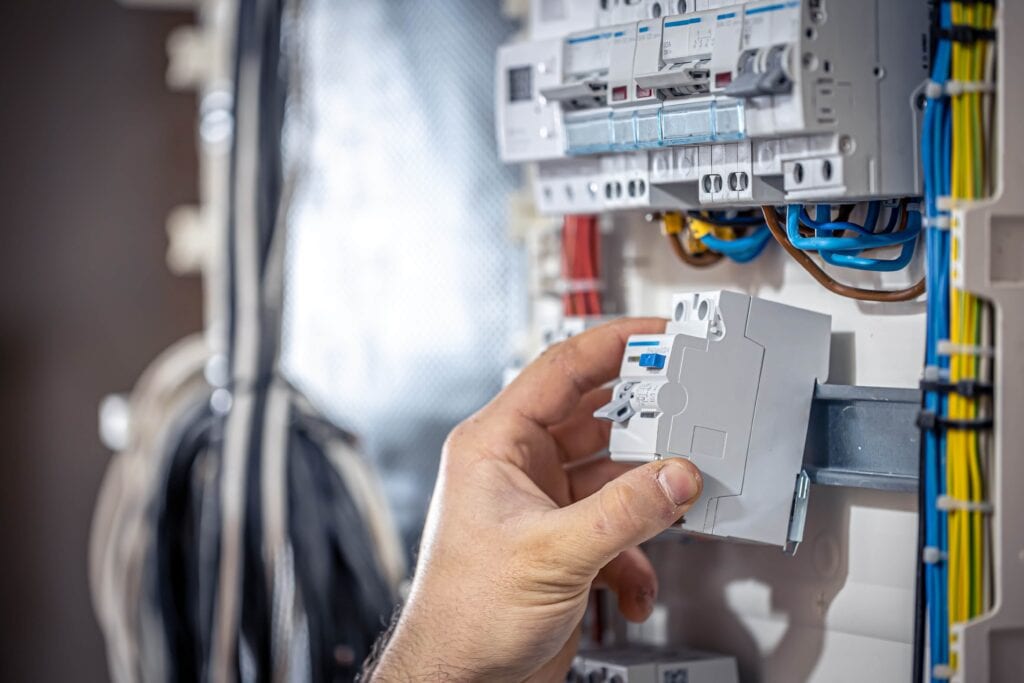 A close-up of a male electrician's hand installing or replacing a white circuit breaker into a residential electrical switchboard with colorful wiring.