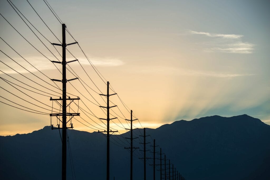 A row of tall wooden utility poles and electrical power lines silhouetted against a mountain range and a glowing golden sunset.