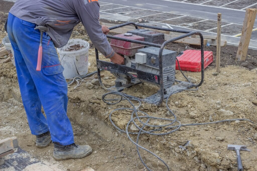 A construction worker in blue work pants and a grey jacket starting a portable electric generator on a dirt construction site.