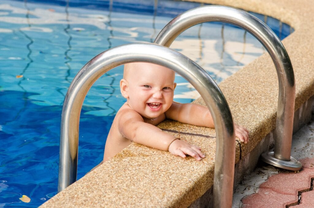 A happy, laughing baby with a wide smile holding onto the edge of a blue swimming pool next to metal handrails.