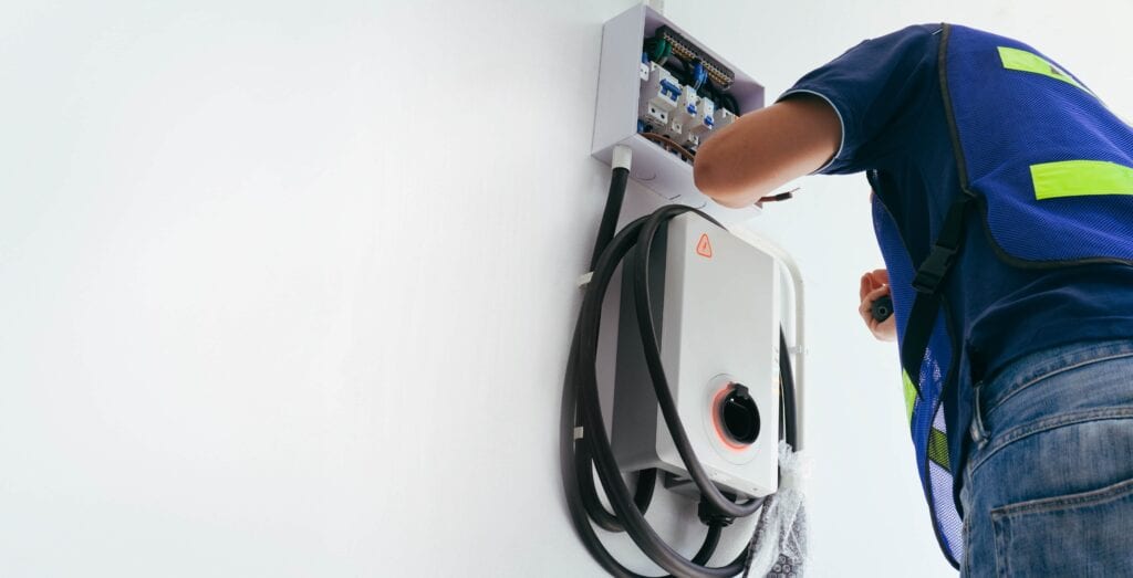 A certified male electrician in a blue safety vest wiring a wall-mounted electric vehicle (EV) charging station and circuit breaker box on a white garage wall.