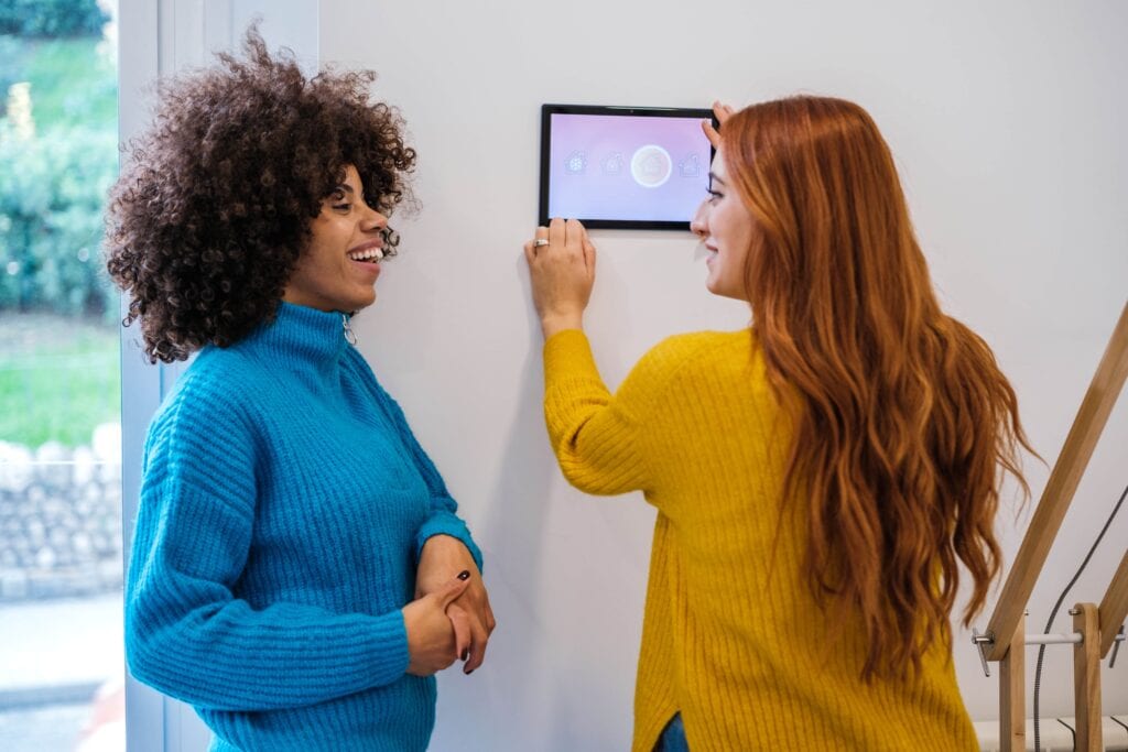 Two women in colorful sweaters—one with curly dark hair and one with long red hair—smiling while interacting with a wall-mounted smart home control tablet in a modern living space.