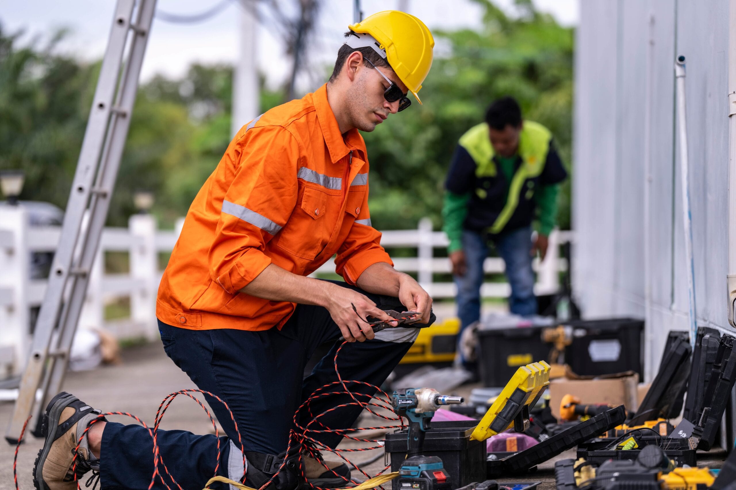 A technician kneeling on the ground wearing a bright orange high-visibility safety jacket, yellow hard hat, and sunglasses, carefully stripping or splicing wires with hand tools surrounded by open toolboxes and power equipment.