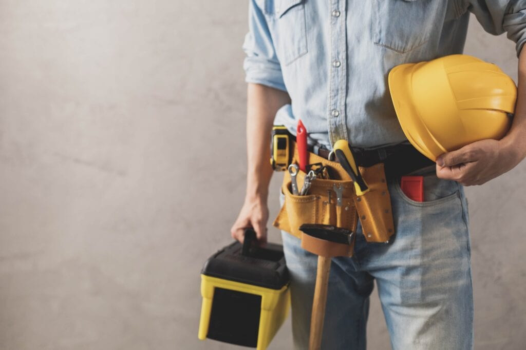 Mid-section view of a construction worker in a denim shirt and jeans, wearing a loaded leather tool belt, carrying a yellow toolbox, and holding a yellow hard hat against a neutral concrete background.