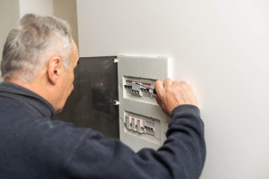 An elderly man with grey hair, seen from behind, using a screwdriver to work on a white electrical fuse box or circuit breaker panel mounted on a plain white wall.
