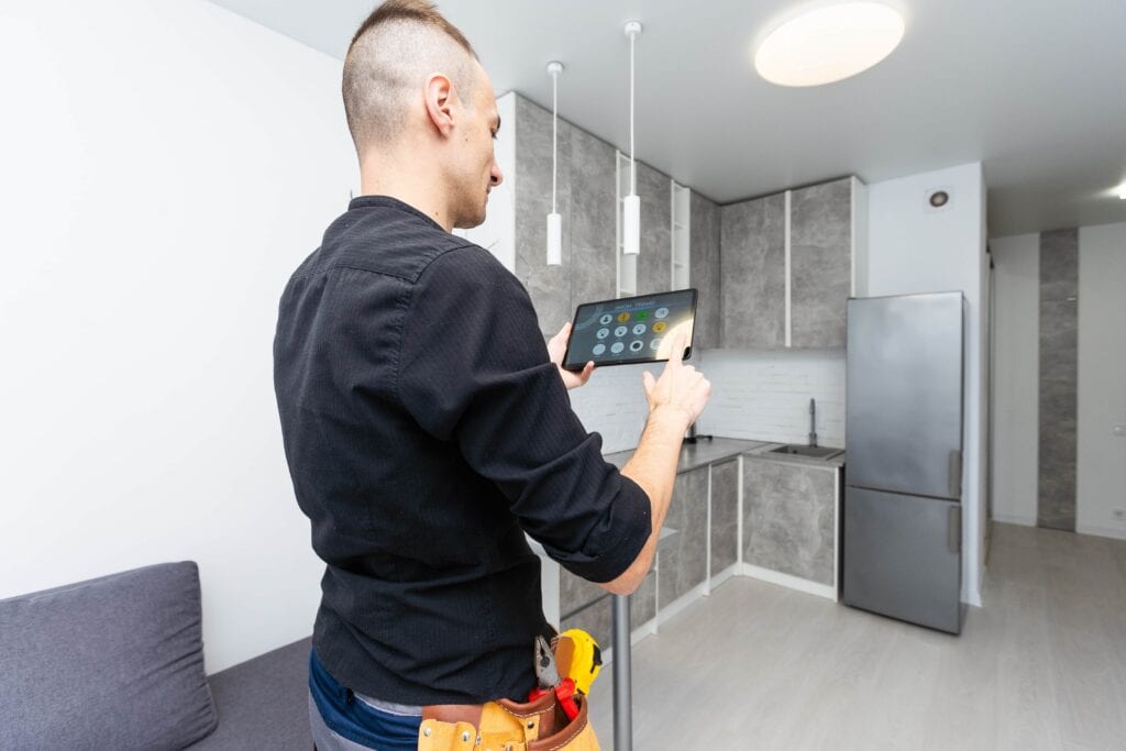 Over-the-shoulder view of a technician wearing a tool belt while using a digital tablet to control a smart home interface in a modern gray and white kitchen.
