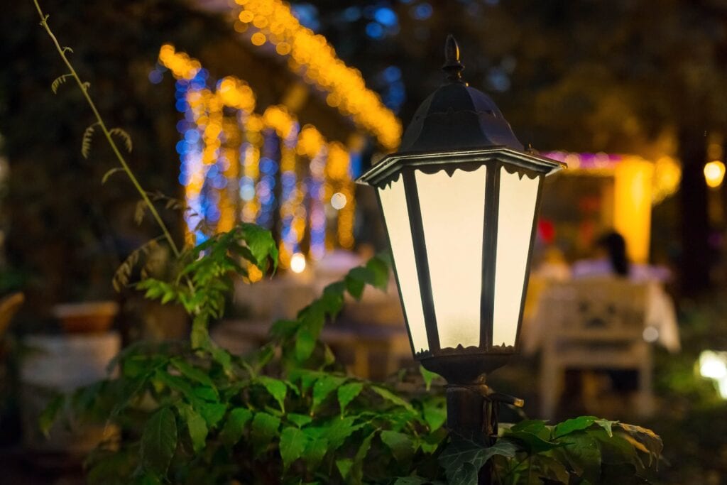 A black iron retro-style garden lantern glowing with warm light in the foreground, with lush green leaves below and a soft bokeh background of golden fairy lights in a night garden setting.