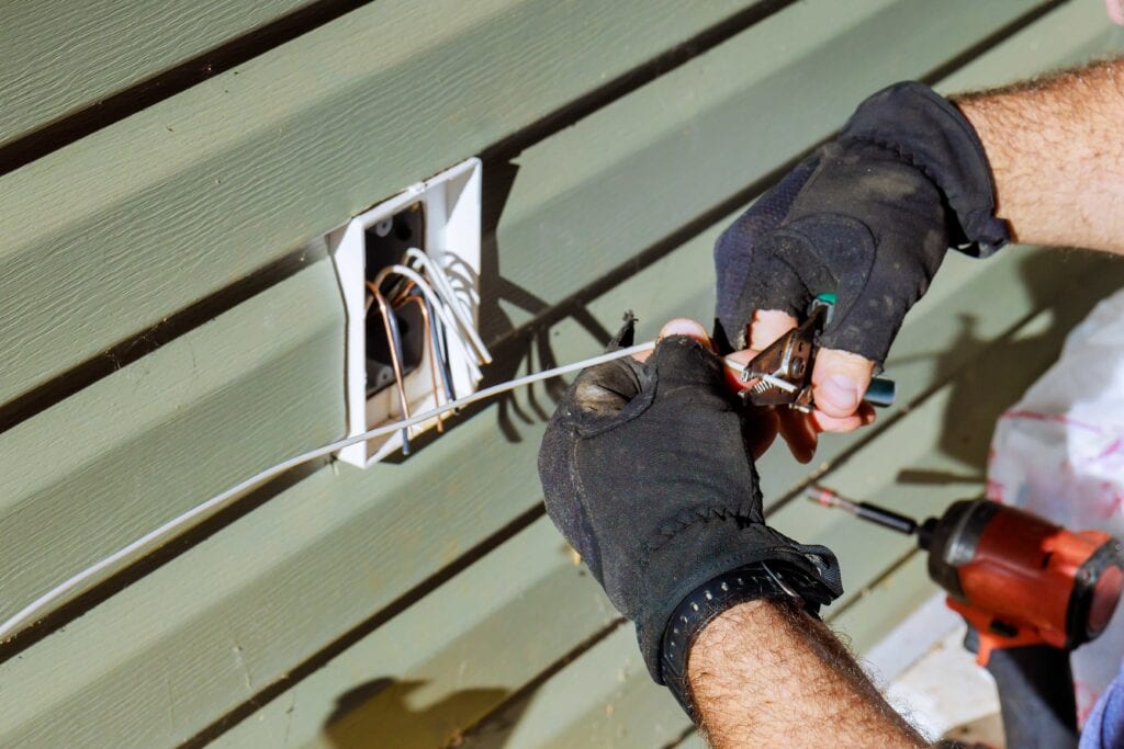 A licensed electrician wearing protective gloves uses wire strippers to prepare exterior wiring for a new outdoor outlet installation in Florida.